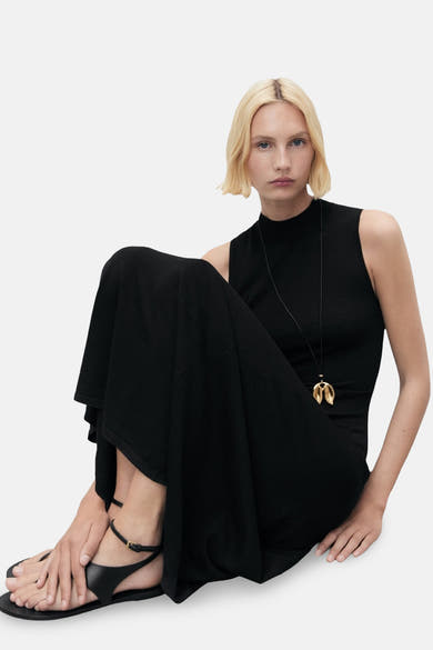 Zwarte nauwsluitende jurk van MANGO, met een opstaande kraag en een lange uitlopende rok, gedragen met een gouden ketting en platte sandalen.