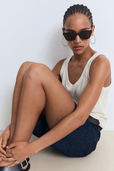 Beige tanktop van MANGO, gedragen met een zonnebril en gouden oorringen, zittend op de grond.