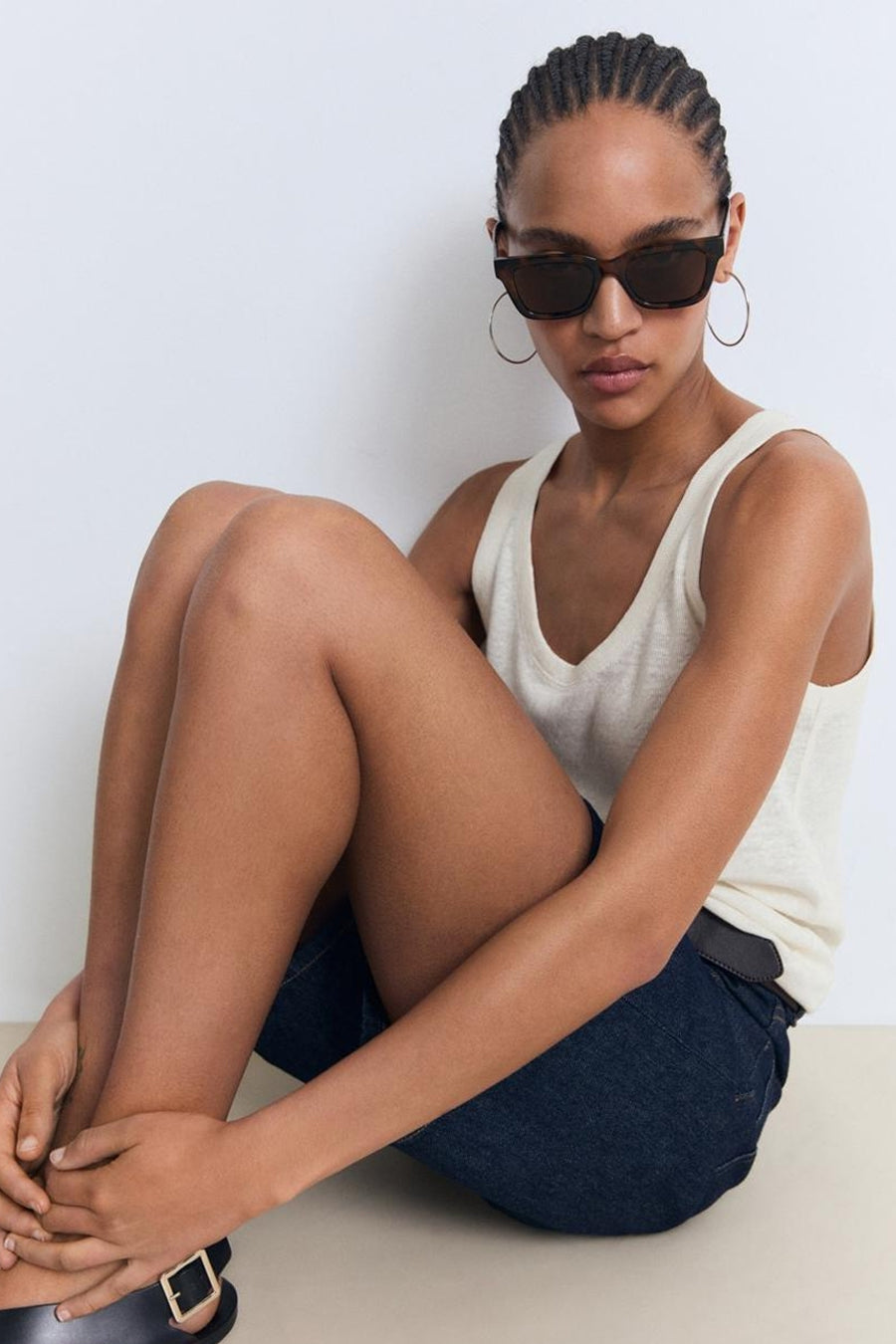 Beige tanktop van MANGO, gedragen met een zonnebril en gouden oorringen, zittend op de grond.