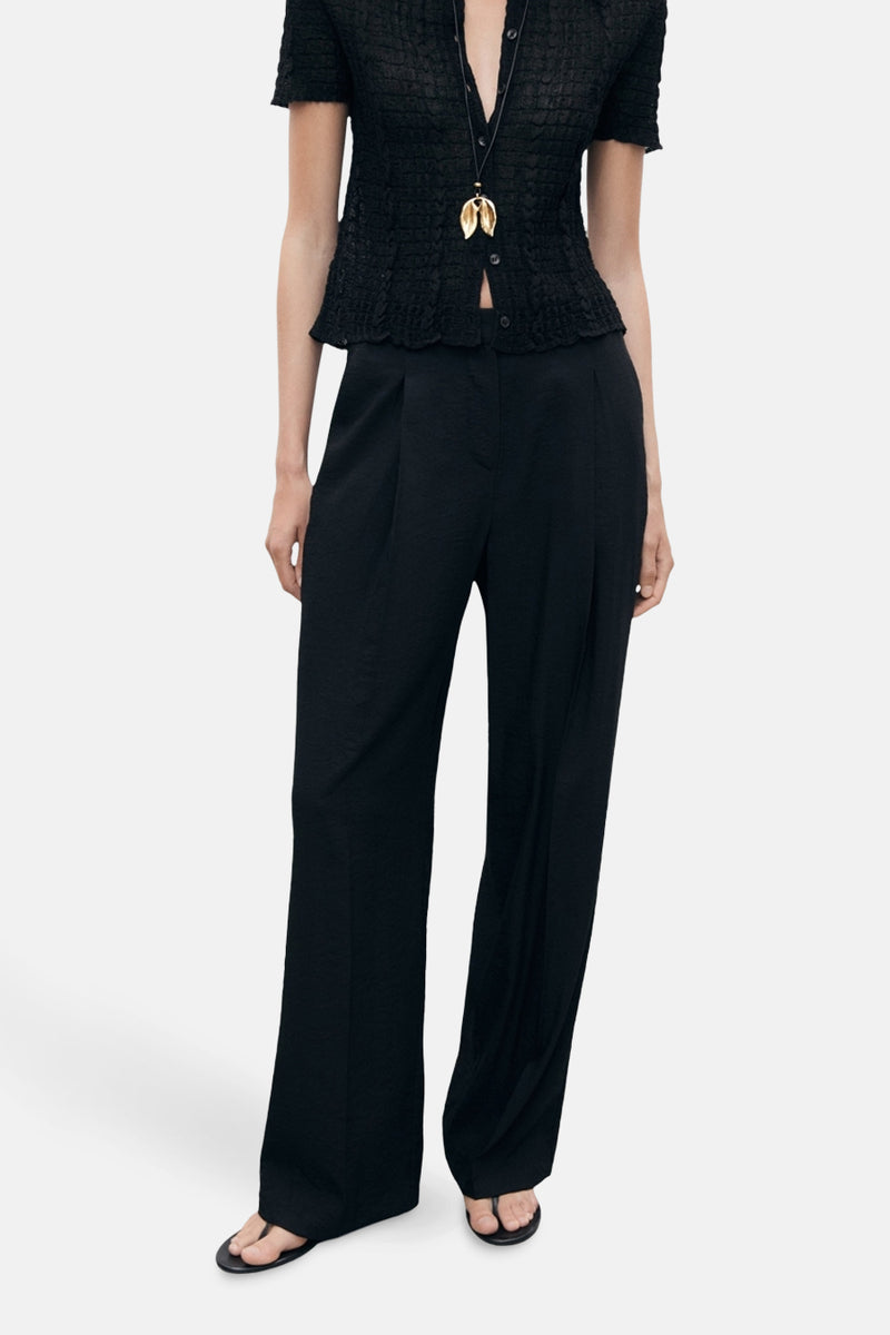 Zwarte broek van MANGO, losse pasvorm met vouwen en plooien aan de voorkant, gedragen met een top en een ketting.