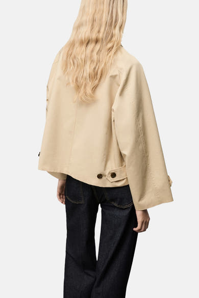 Veste courte beige de MANGO, avec manches amples et détails de boutons au dos, portée avec un jean foncé.