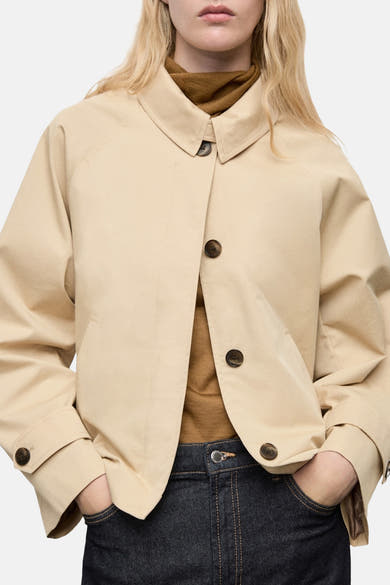 Veste courte beige de MANGO, boutonnée avec un col classique, portée avec un pull marron et un jean bleu foncé.