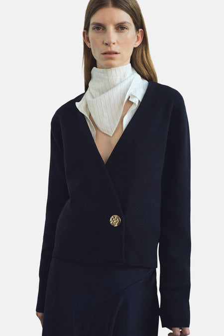 Blauwe MANGO-gilet, gouden knoop, gedragen over een wit gestreept overhemd met een opstaande kraag.