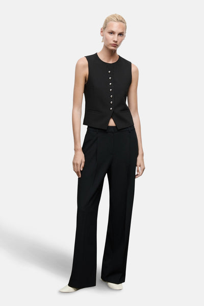 Pantalon noir de MANGO, coupe droite avec des plis, porté avec un gilet noir boutonné et des chaussures blanches.