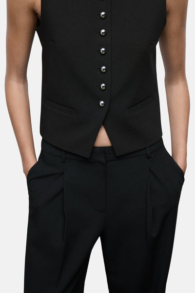 Pantalon noir de MANGO, avec des plis et poches latérales, porté avec un gilet noir boutonné.
