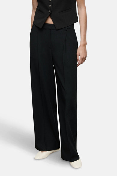 Pantalon noir coupe droite par MANGO, avec plis et poches latérales, porté avec un gilet noir et des chaussures blanches.
