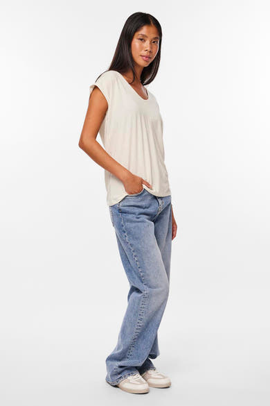Wit T-shirt met korte mouwen van PIECES®, gedragen met een wijde blauwe spijkerbroek en crèmekleurige sneakers, zijwaarts gezien.