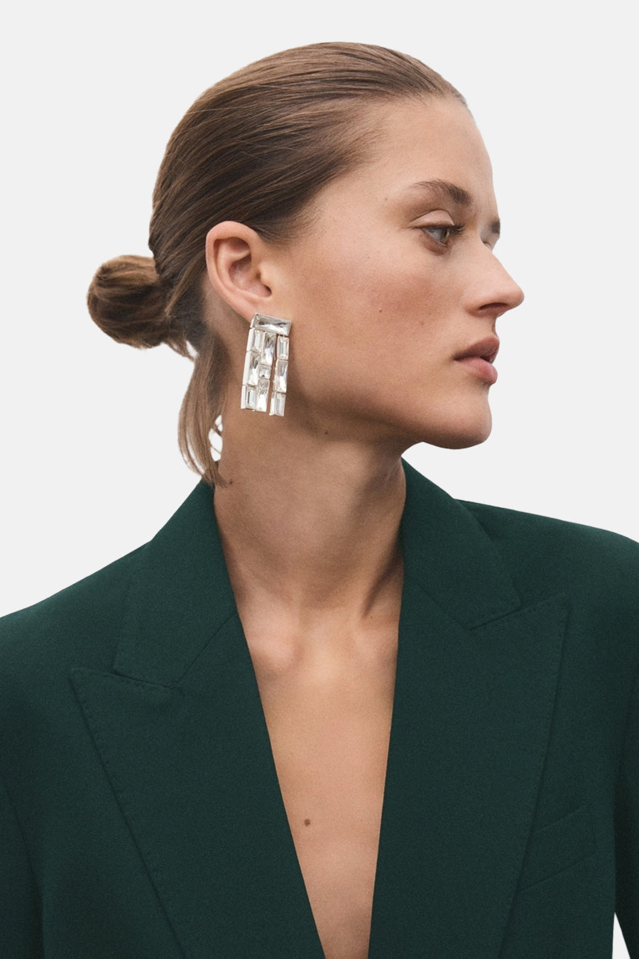 Groene blazer van MANGO, gedragen met rechthoekige hangende oorbellen met transparante stenen en het haar opgestoken in een knot.