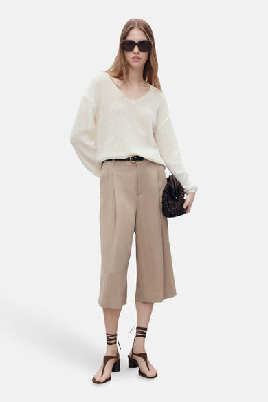Pull à col en V blanc de MANGO, porté avec un pantacourt beige et des sandales à lacets marron.
