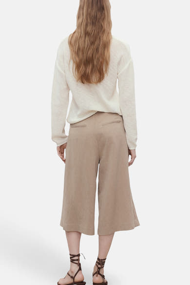 Pull à col en V blanc de MANGO, porté avec un pantalon culotte beige et des sandales à lanières.