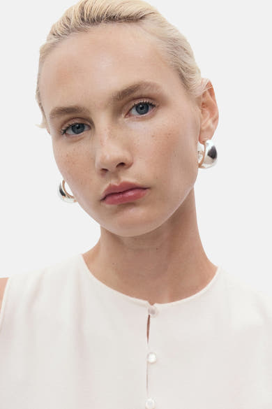 Gilet blanc de MANGO avec une patte de boutonnage nacrée, porté avec des boucles d'oreilles argentées.
