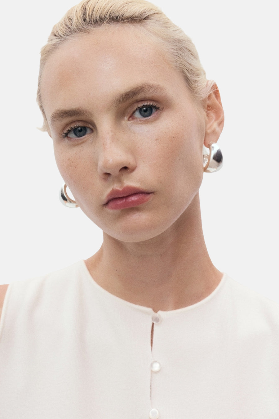 Gilet blanc de MANGO avec une patte de boutonnage nacrée, porté avec des boucles d'oreilles argentées.
