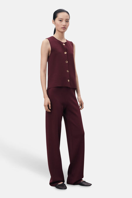 Blouse bordeaux sans manches de MANGO, avec boutons dorés, portée avec un pantalon assorti et des ballerines noires.