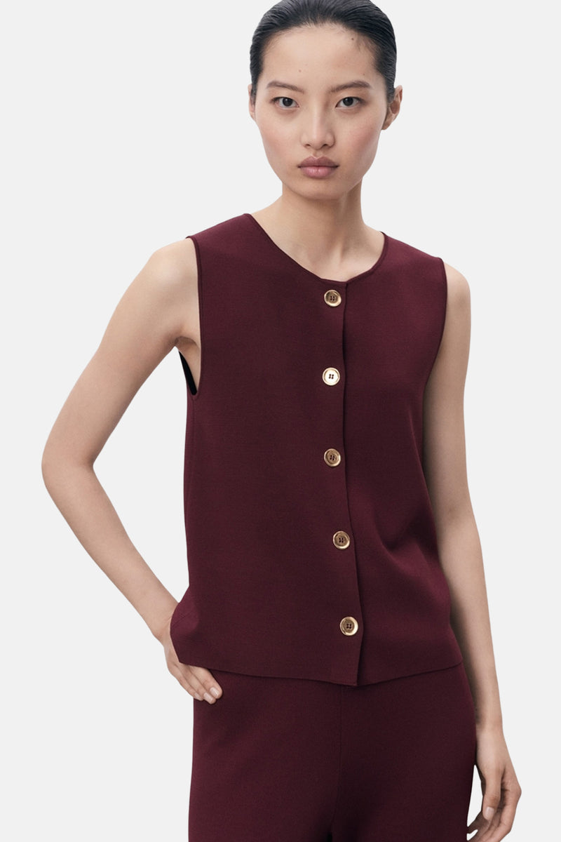 Blouse bordeaux sans manches de MANGO, fermée par des boutons dorés, portée avec un pantalon bordeaux.