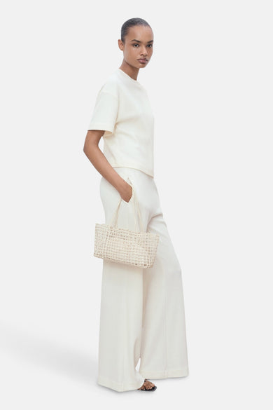 Pantalon blanc de MANGO, ample et taille haute, porté avec un t-shirt crème et un sac à main en osier.
