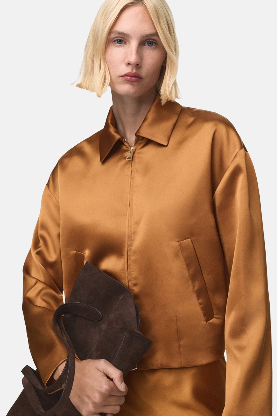 Gilet marron de MANGO avec une texture satinée et un sac à main en daim dans la main.