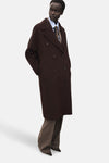 Veste longue brune par MANGO, croisée avec des boutons, portée avec une chemise bleu clair et un pantalon à carreaux marron.