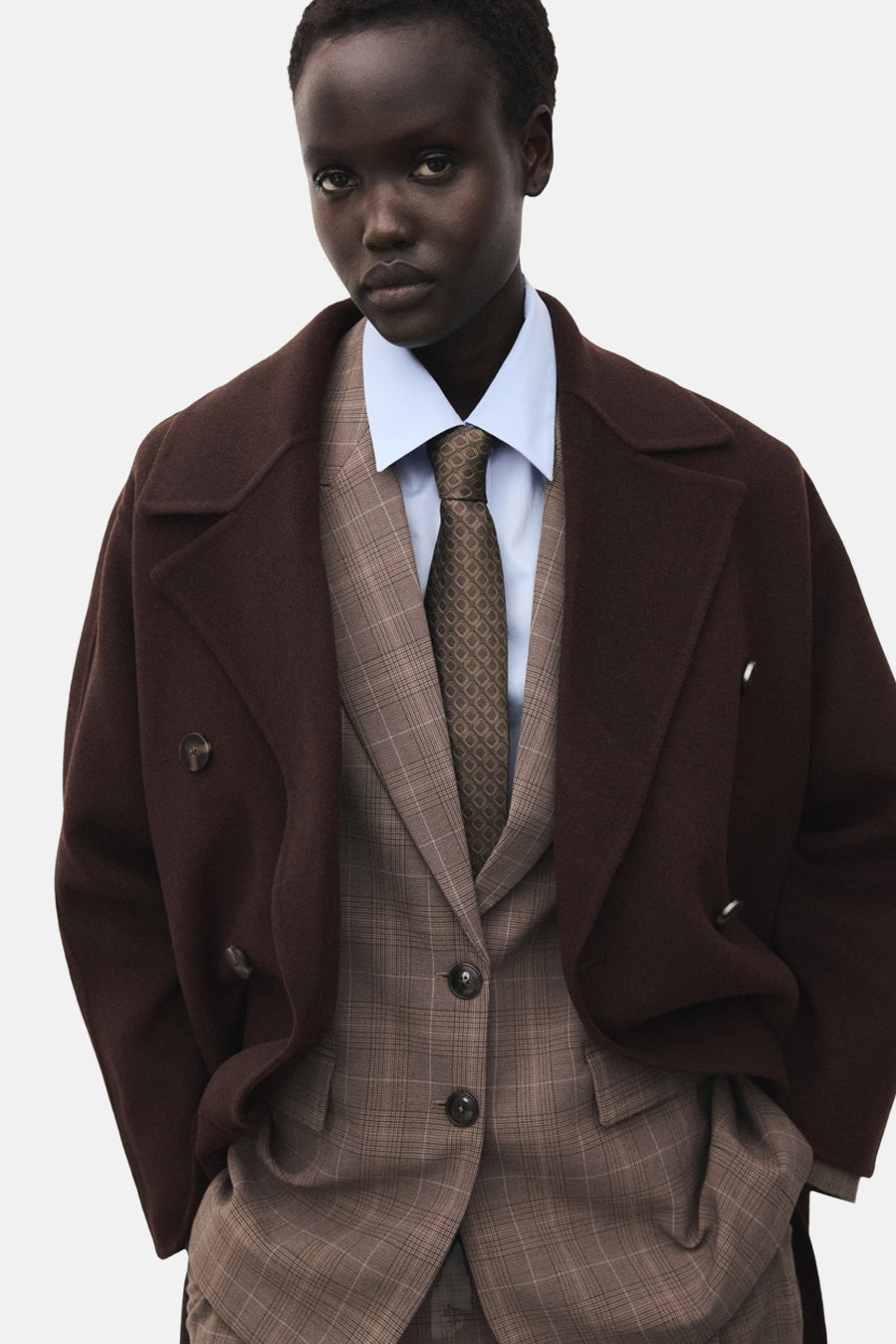 Veste longue marron de MANGO, croisée, portée sur un tailleur à carreaux avec chemise bleu clair et cravate marron.