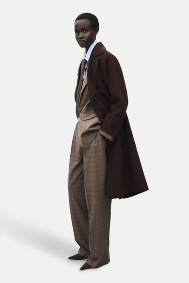 Veste longue brun de MANGO, portée sur un costume à carreaux avec une chemise bleu clair et une cravate.