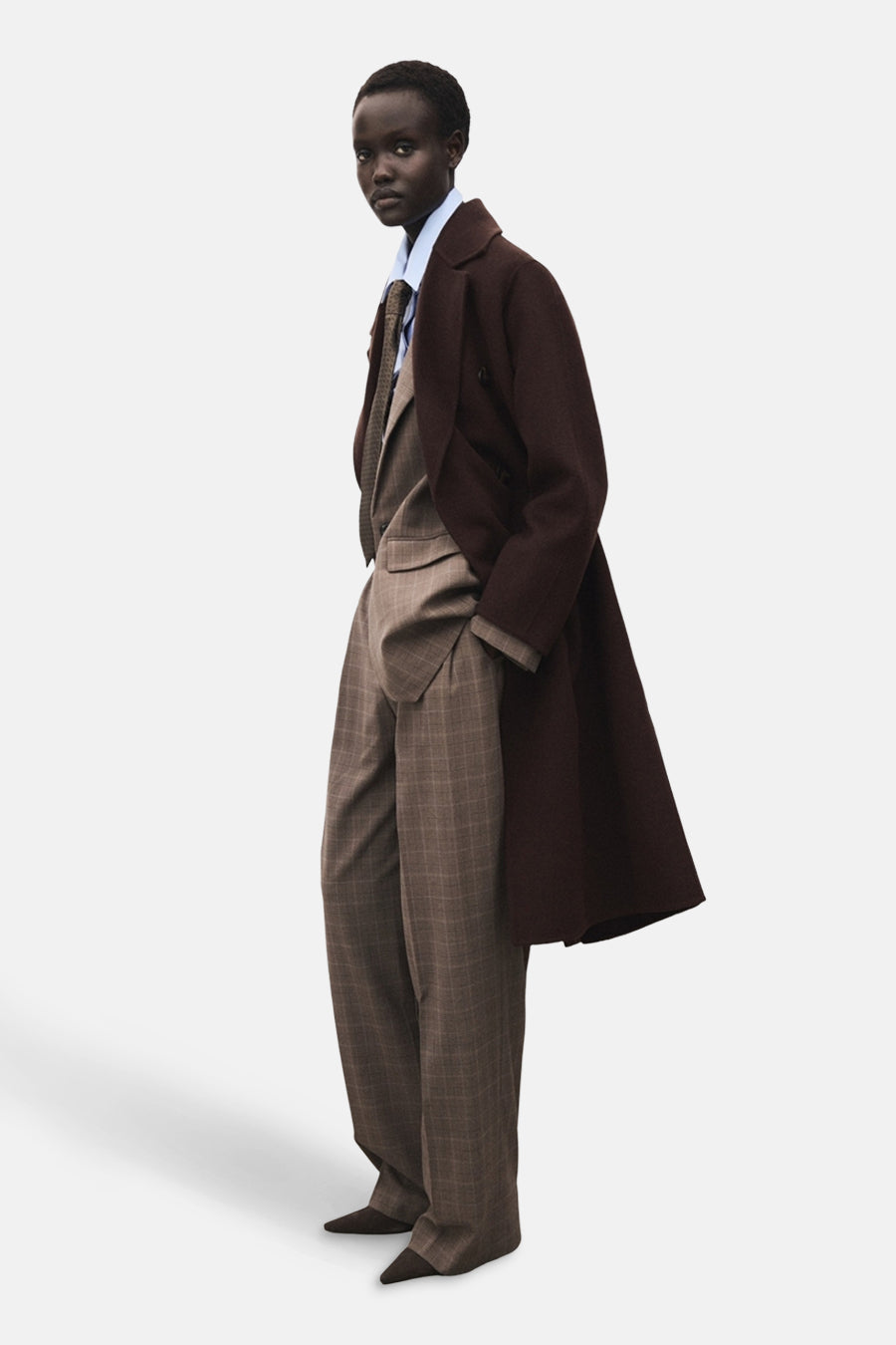 Veste longue brun de MANGO, portée sur un costume à carreaux avec une chemise bleu clair et une cravate.