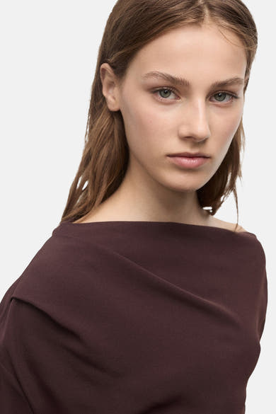 Bruine blouse met lange mouwen van MANGO, asymmetrische snit die de schouder accentueert, op een effen achtergrond.