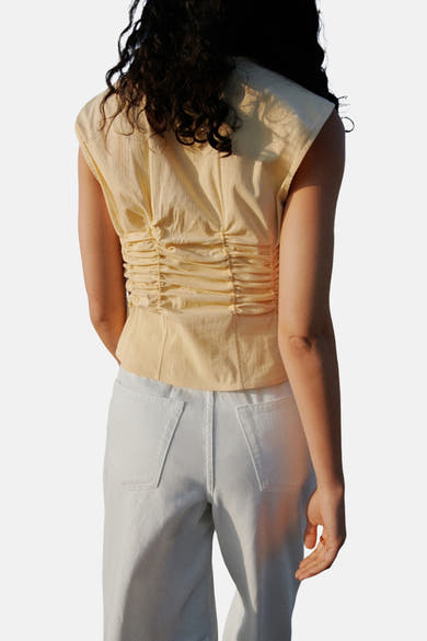 Gele mouwloze blouse van MANGO, met geplooide details op de rug en gedragen met een witte broek.