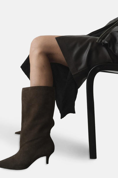 Bottes marron en daim de MANGO, portées avec une jupe noire en cuir, assise sur une chaise noire, vue rapprochée.
