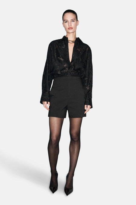 Chemisier noir à manches longues de MANGO, avec motif floral ton sur ton, porté avec des shorts taille haute noirs et des collants.
