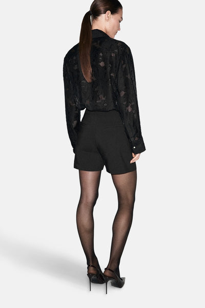 Chemise noire à manches longues de MANGO, avec motif floral en ton sur ton, portée avec un short noir et des collants noirs.