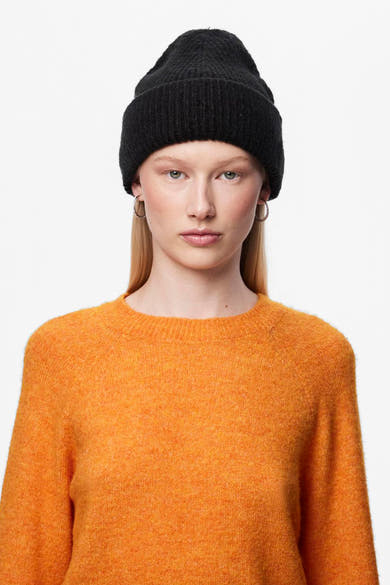 Bonnet noir de PIECES, porté par une femme aux cheveux blonds et aux boucles d'oreilles argentées, avec un pull orange.