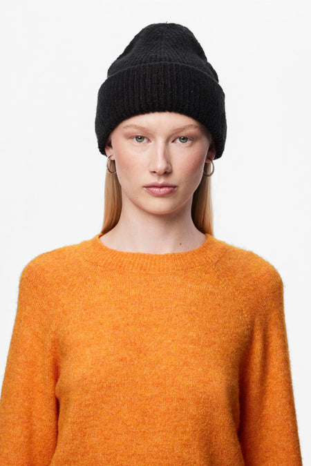Zwarte muts van PIECES, gedragen door een vrouw met blond haar en zilveren oorbellen, met een oranje trui.