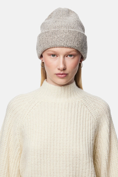 Bonnet beige en tricot par PIECES®, porté avec des boucles d'oreilles argentées et un pull blanc à col roulé.