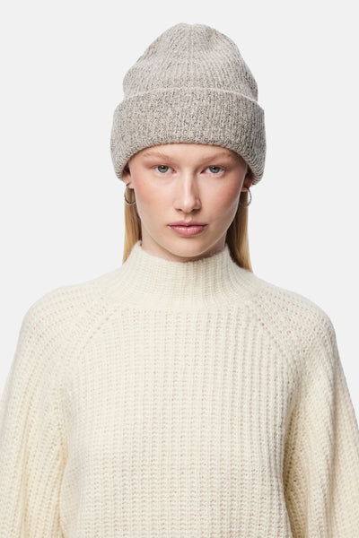 Bonnet beige en tricot par PIECES®, porté avec des boucles d'oreilles argentées et un pull blanc à col roulé.