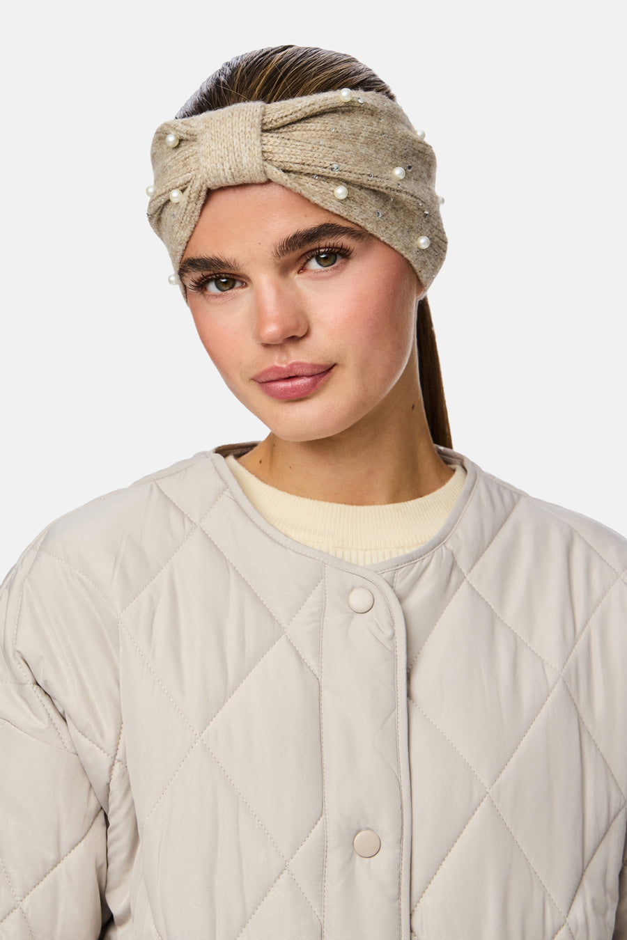 Chauffe-oreille beige de PIECES® avec un nœud et des perles décoratives, porté avec une veste matelassée.
