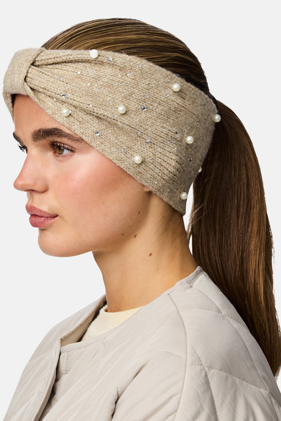 Chauffe-oreille beige de PIECES®, orné de perles et de strass, porté avec une veste matelassée claire.
