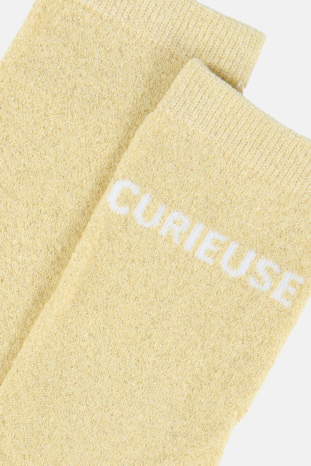 Chaussettes jaunes scintillantes de PIECES® en maille chevron avec l'inscription blanche 'CURIEUSE'.