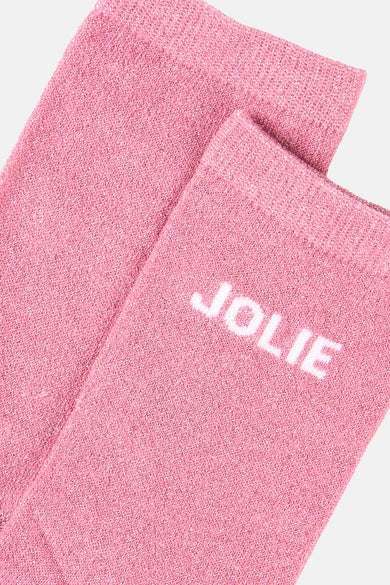 Chaussettes roses scintillantes de PIECES, avec le mot « JOLIE » en blanc, sur fond uni.