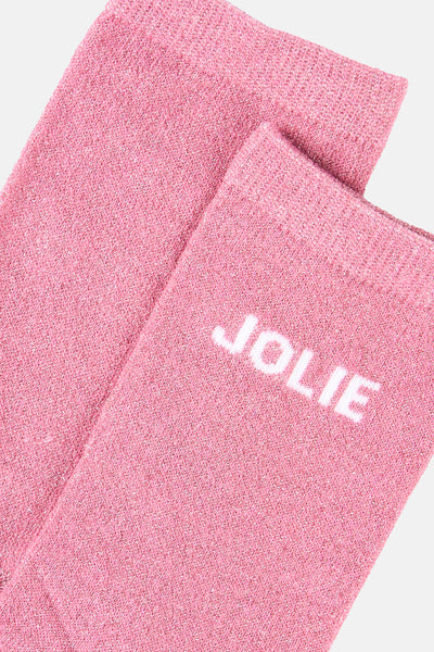 Chaussettes roses scintillantes de PIECES, avec le mot « JOLIE » en blanc, sur fond uni.