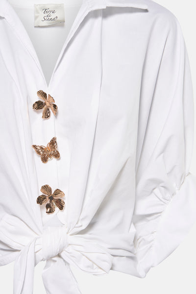 Chemisier blanc à manches courtes par Terra di Siena, avec boutons décoratifs floraux et papillon, et un nœud à la taille.
