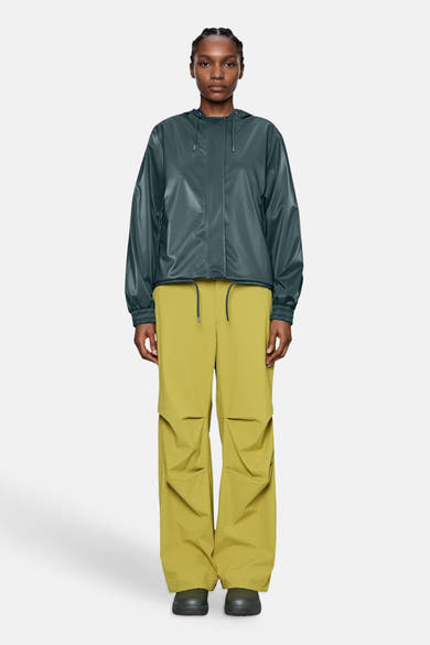 Dark green shiny casual jacket with hood and elastic cuffs, worn with mustard cargo pants and black shoes, front view