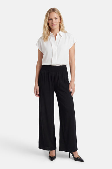 Black wide-leg elastic waist trousers with pleated front, worn with white blouse and black heels, full-length front view