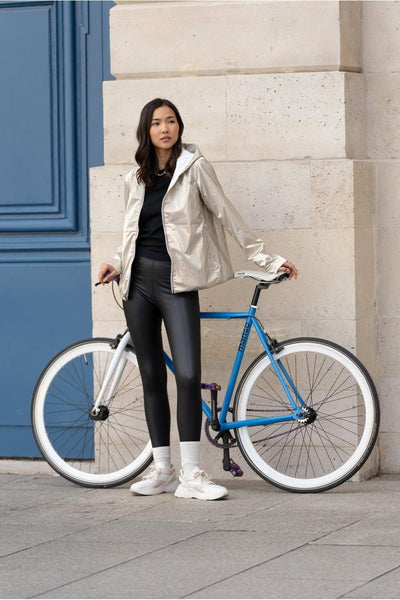 Veste de pluie écru de LAURA JO, portée avec un t-shirt noir, un legging en cuir, des baskets et un vélo bleu.