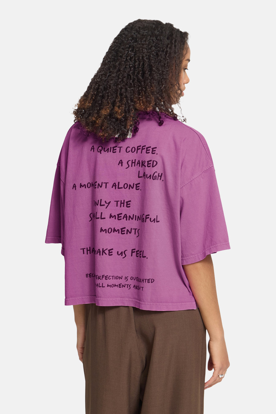 Rose oversized short-sleeve t-shirt with black handwritten text print, back view, worn with brown wide-leg pants