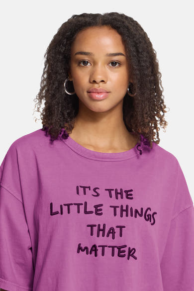 Rose short-sleeve t-shirt with embroidered text "IT'S THE LITTLE THINGS THAT MATTER" in dark purple, worn by model with curly hair and hoop earrings, front view.