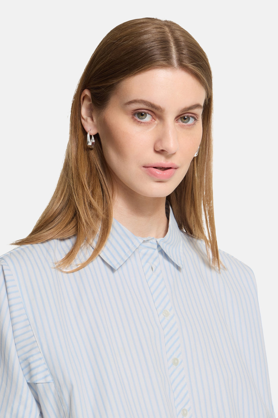 Blue and white striped long-sleeve blouse with pointed collar and front buttons, shown in close-up on model with straight brown hair and silver hoop earrings.