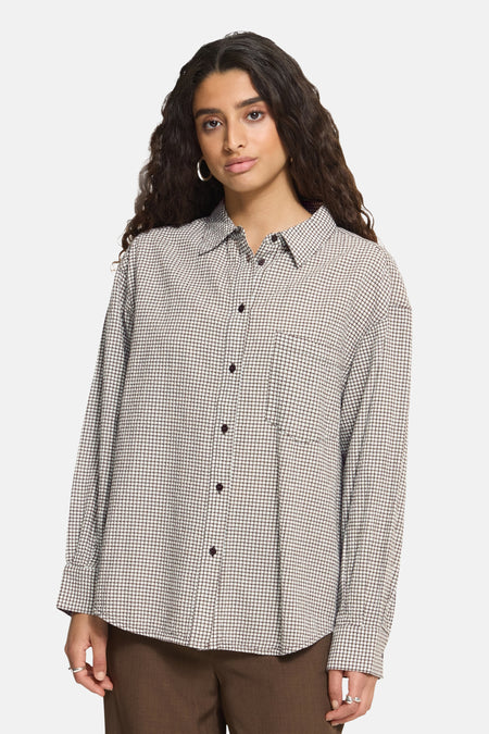 Multicolored long-sleeve checkered chemisier by ICHI, featuring front buttons and chest pocket, worn with brown pants and silver rings.