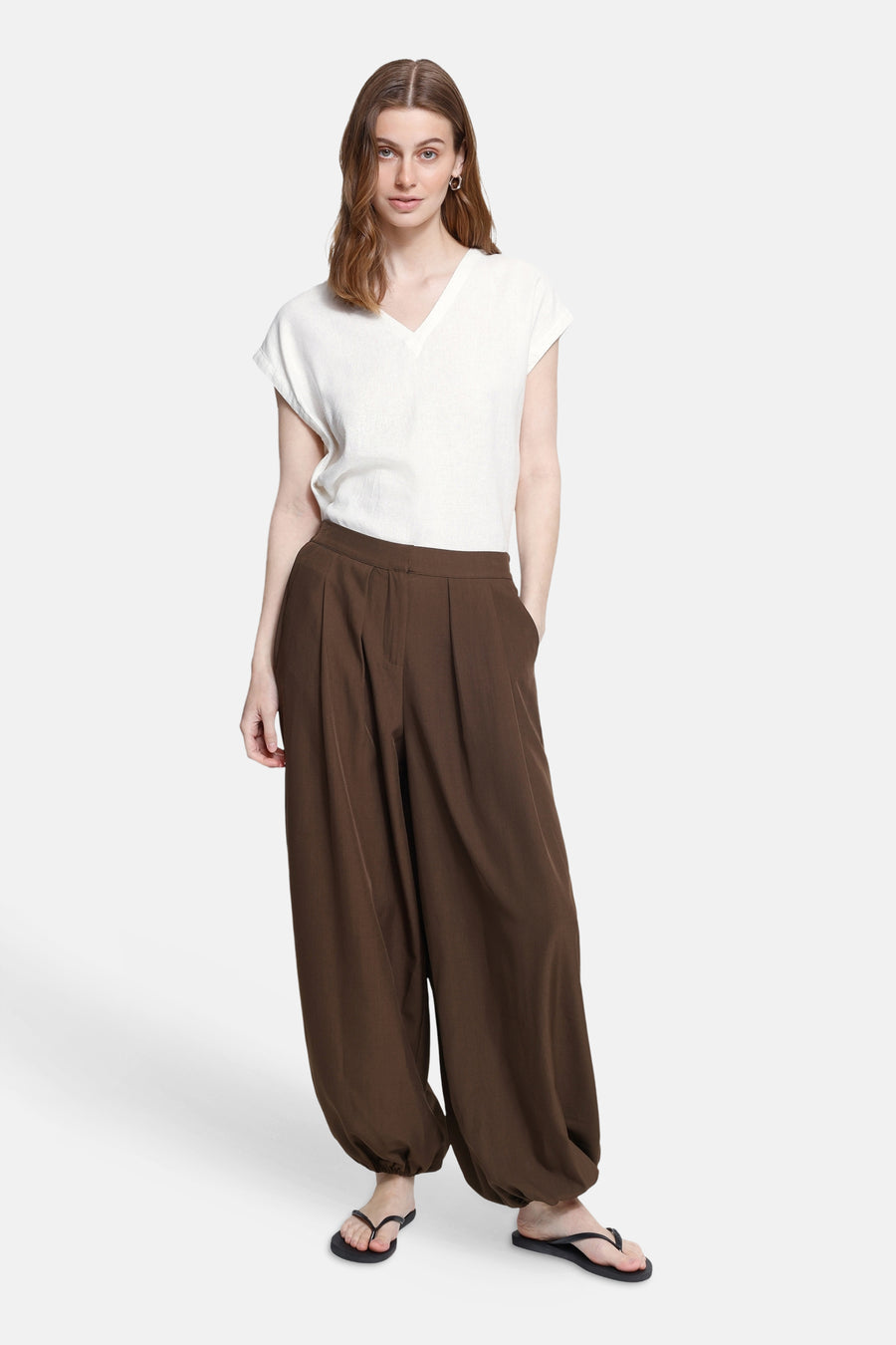 Brown wide-leg elastic waist trousers with pleated front and gathered ankles, worn with white top and black flip-flops, full-length front view