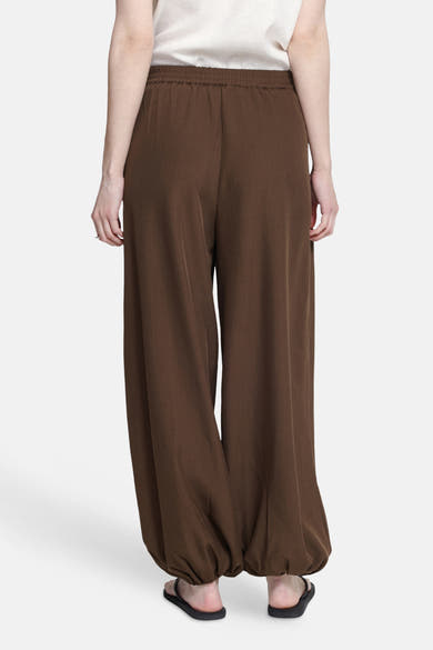 Brown elastic-waist wide-leg trousers with gathered ankle cuffs, worn with a white top and black sandals, back view.