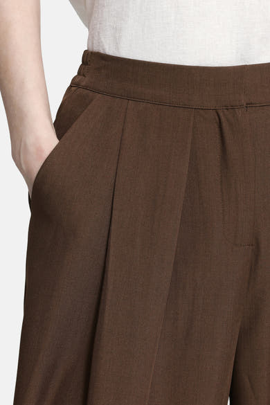 Brown elastic-waist women's seasonal trousers with front pleats and side pockets, worn with a light gray top, close-up view