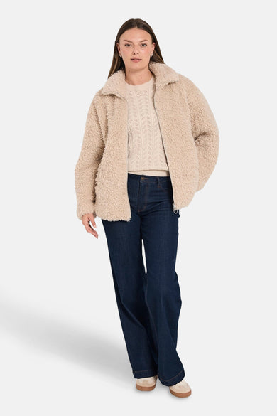 Veste courte beige en fausse fourrure de FREEQUENT, portée avec un pull en tricot à torsades et un jean évasé.
